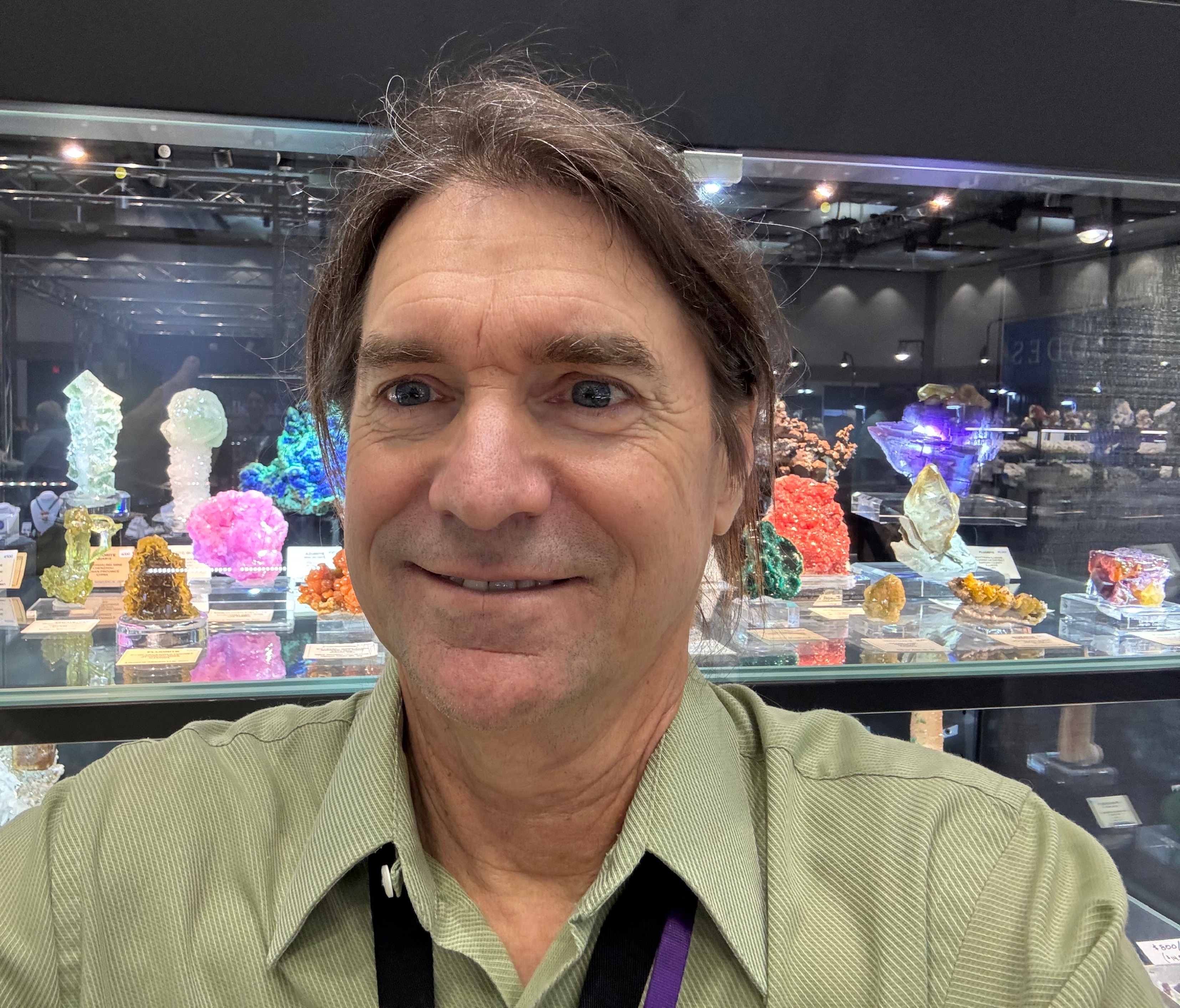 A photograph in color of Evan Jones, shoulder and up. He is wearing a green colored shirt and his brown hair, speckled with gray, is longer and pulled back. He smiles into the camera and stands in front of more than a dozen, very colorful mineral specimesn in a glass case, where one shelf can be seen behind him.